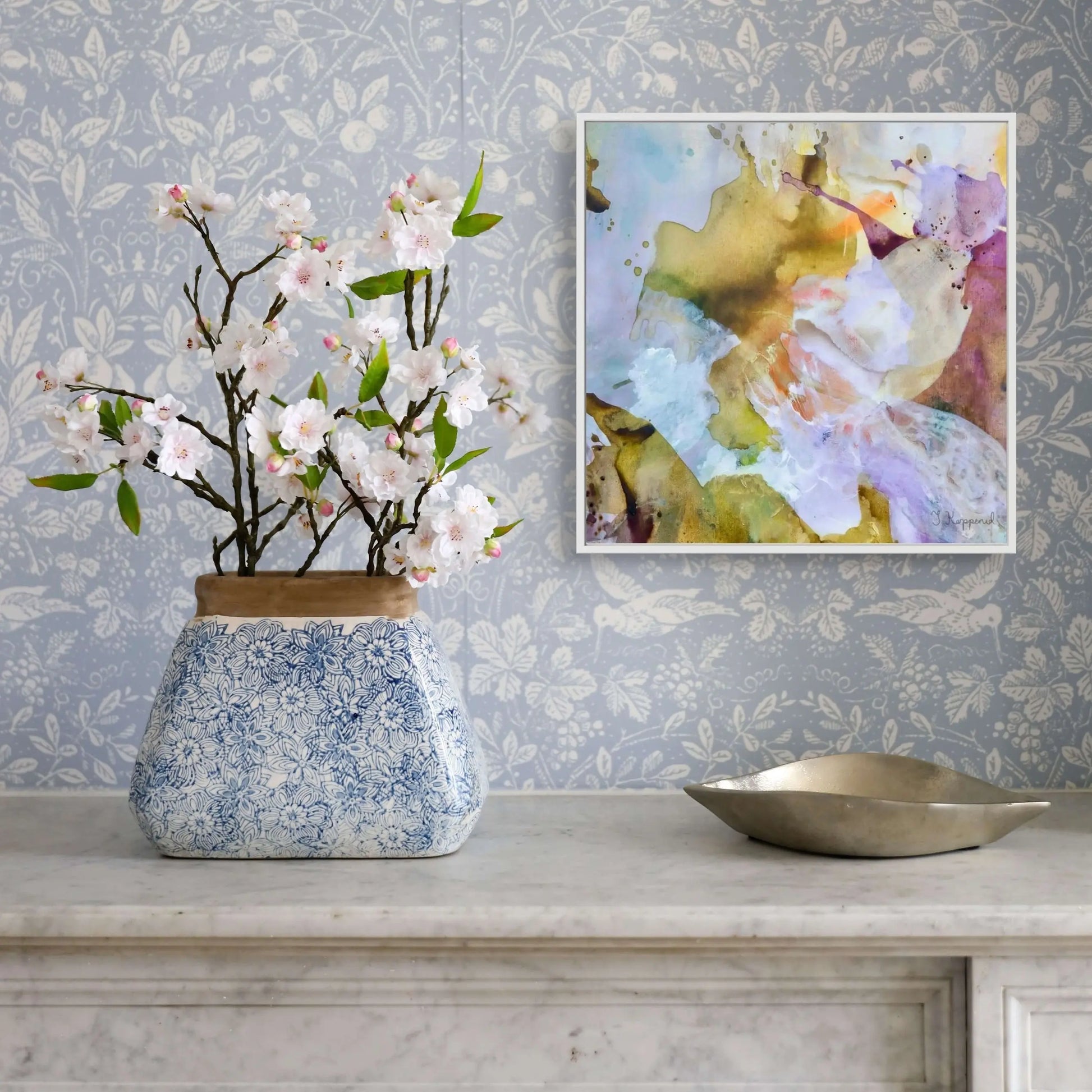Decorative vase with flowers on a table against a patterned wall with a framed abstract painting.